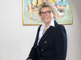 Viviana Cattaneo, CEO di Saturn Over Srl, ritratta in un ambiente professionale elegante. Indossa un completo scuro con bottoni dorati e camicia bianca, seduta davanti a una parete chiara con un quadro astratto dai colori vivaci incorniciato in oro. L’immagine trasmette leadership, professionalità e attenzione ai dettagli, in linea con il ruolo di guida di una boutique dell’outsourcing innovativa.