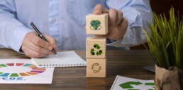 Mano che impila cubi di legno con icone di sostenibilità (cuore con il pianeta, riciclo, economia circolare) su una scrivania con documenti dell’Agenda 2030 e una pianta verde, rappresentando la sintesi di sostenibilità di Saturn Over Srl