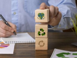 Mano che impila cubi di legno con icone di sostenibilità (cuore con il pianeta, riciclo, economia circolare) su una scrivania con documenti dell’Agenda 2030 e una pianta verde, rappresentando la sintesi di sostenibilità di Saturn Over Srl
